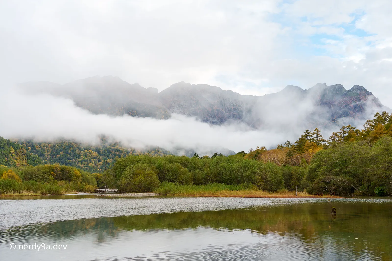 Taisho Pond in Kamikochi reflecting vibrant autumn forests with the rugged peaks of the Hotaka mountain range shrouded in low-hanging clouds and mist
