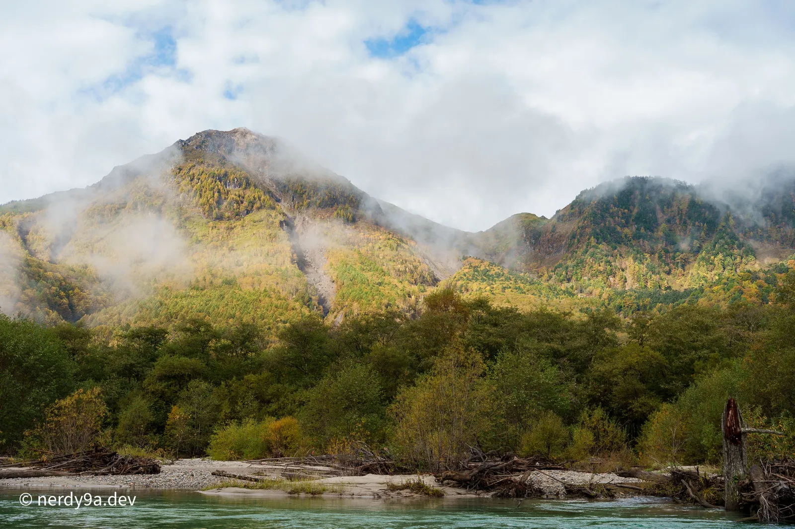 The Azusa River flowing with driftwood on its gravel bank under a cloud-shrouded Mt. Yakedake in Kamikochi
