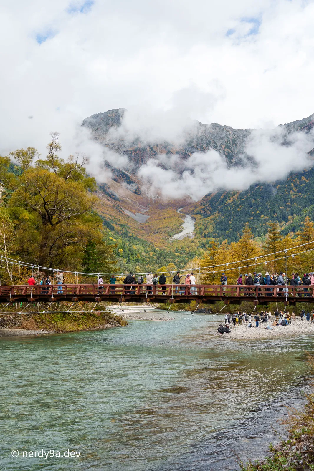 Wide portrait view of Kappa Bridge packed with tourists, spanning the clear turquoise Azusa River with the dramatic, cloud-shrouded peaks of Mount Hotaka in the background