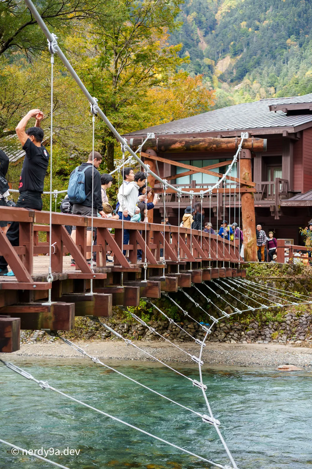 Close-up side view of the wooden structure and suspension cables of Kappa Bridge with tourists standing on the deck and a wooden lodge in the background