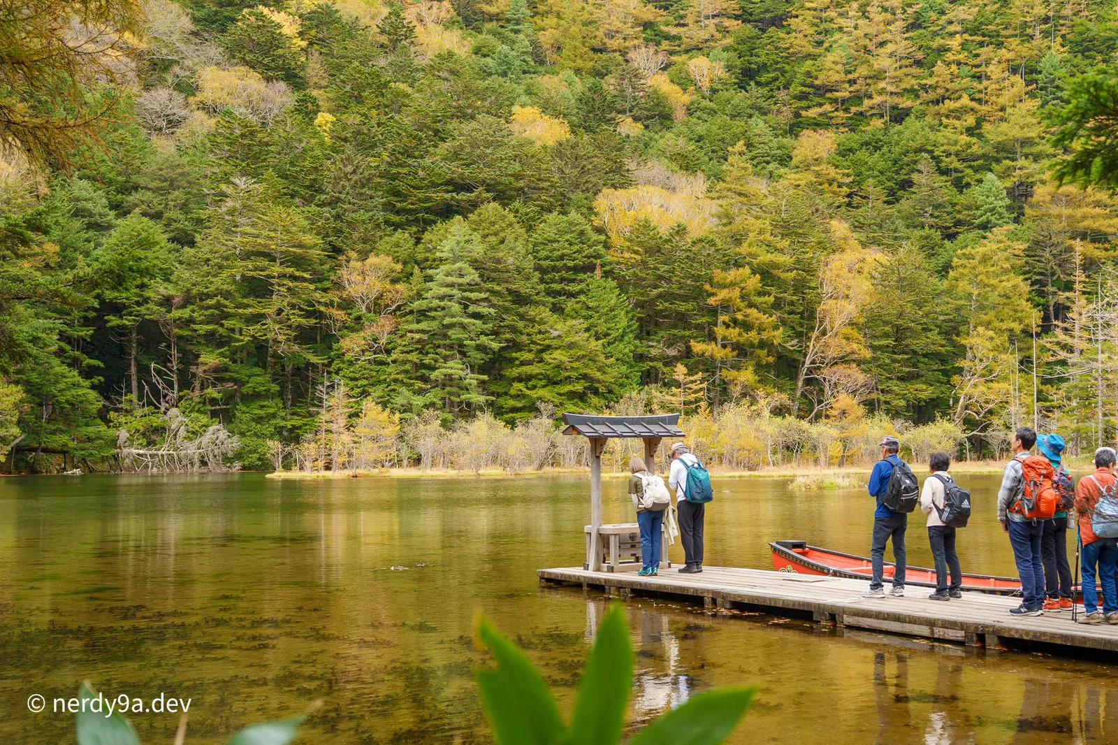 Tourists standing on a wooden pier with a small wooden altar and a red boat at Myojin Pond, set against a forested mountain background