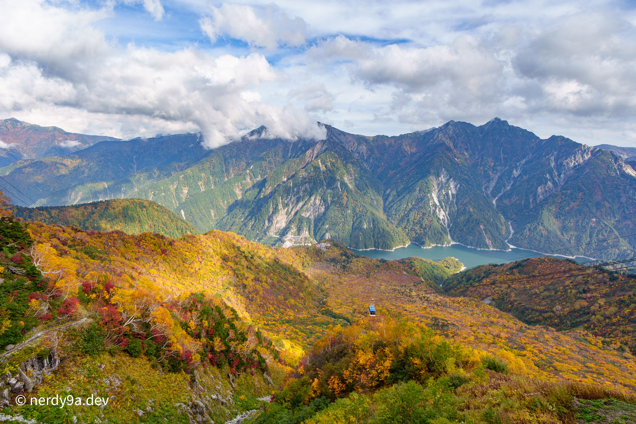 Panoramic view from Tateyama Ropeway showing a cable car cabin suspended over a valley filled with autumn foliage, looking towards Kurobe Lake