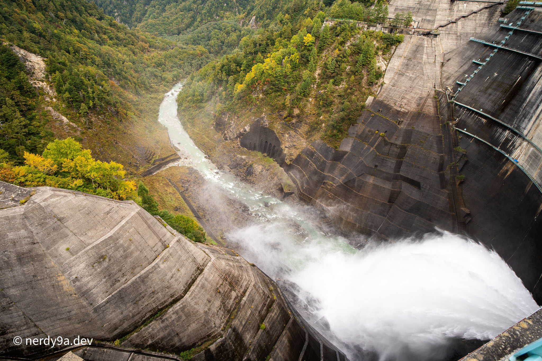 High-angle view of massive water discharge spraying from Kurobe Dam into the green river valley below